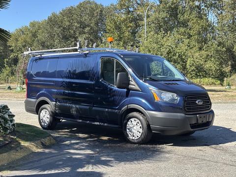 A blue 2016 Ford Transit 150 van with a roof rack and orange beacon light on top parked with its side facing the viewer