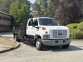 A white 2005 GMC C7500 Topkick truck with a flatbed and black side rails parked on a road with a clear view of the front and side of the vehicle