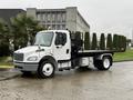 A 2019 Freightliner M2 flatbed truck with a white cab and black flatbed parked at an angle with a black grille and lights on top