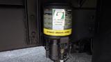 A close-up view of an automatic greasing system attached to a 2003 International 5600i with a gray cylindrical container and technical information labels