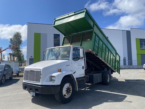 A white 1997 Freightliner FL70 truck with a green dump bed raised in the air for unloading materials