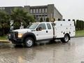 A 2015 Ford F-450 truck with a white utility body and tool compartments on the sides is parked on a wet surface