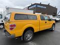 A yellow 2014 Ford F-150 with a cap on the bed and chrome wheels parked at a commercial location