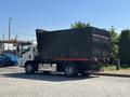 A 2016 Isuzu NPR-HD truck with a large black garbage container attached to the rear and orange accents on the side