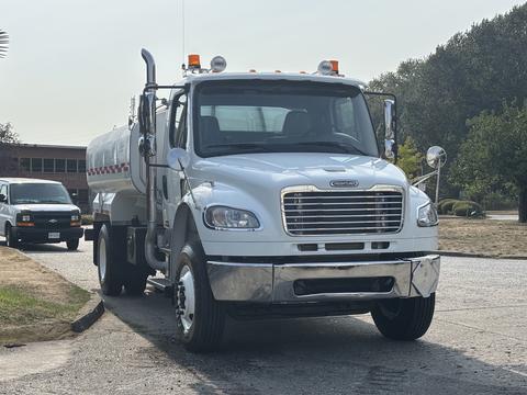 A white 2010 Freightliner M2 106 truck with a tanker attached is parked facing forward with lights on its roof