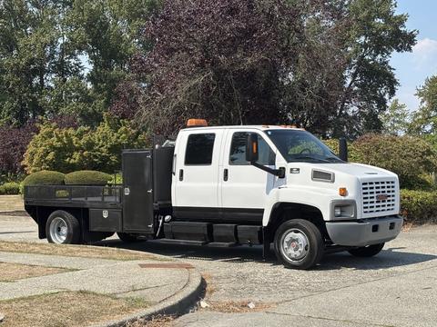 A white 2005 GMC C7500 Topkick truck with a flatbed and an orange light on the roof parked with a tool compartment on the side