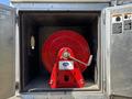 A red hose reel mounted inside a metal storage compartment with a crank handle and a white label on the front