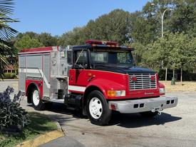 A red 1991 International 4600 fire truck featuring a silver ladder and equipment compartment with a light bar on top and front headlights illuminated
