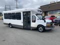 A 2020 Chevrolet Express shuttle bus with a white exterior and large side windows, featuring a front door and signage above the windshield indicating its purpose