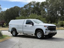 A white 2019 Chevrolet Silverado 1500 with a cap on the bed parked on a concrete surface showcasing its sleek design and chrome accents