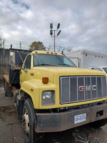 A yellow 1996 GMC C7H042 truck with a flatbed and visible rust on the wheels parked in a yard