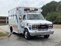 A 1994 GMC Vandura 3500 ambulance with a white body and blue stripes featuring a large light bar on top and an open rear compartment