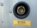 A round metal valve with a golden center and a black rubber gasket affixed to a metallic surface, featuring a yellow informational label about vehicle safety and equipment