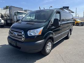 A 2016 Ford Transit van with a dark green exterior and a silver grille parked in a lot with other vehicles in the background