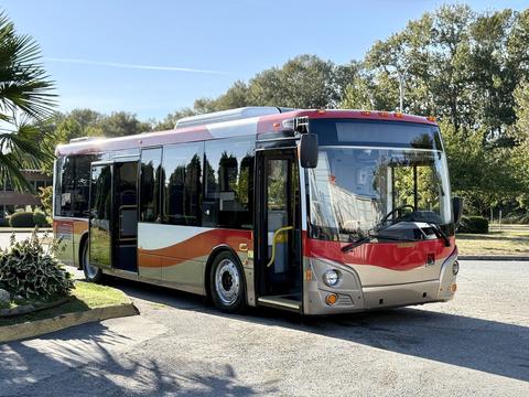 A 2016 Grande West Vicinity bus with a red and silver design parked in a shaded area showcasing its large windshield and modern features