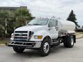 A 2013 Ford F-750 truck with a white tank body and chrome details is parked with its front facing the viewer