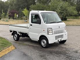 A 2010 Suzuki Carry lightweight truck with a white exterior and a flatbed design is parked facing slightly to the left