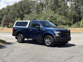 A blue 2015 Ford F-150 with a silver camper shell and chrome wheels parked on a street