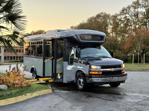 A 2019 Chevrolet Express passenger bus with a black and silver exterior features a ramp for accessibility and is parked at an angle with its doors open