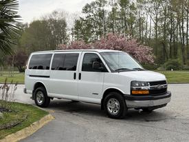 A white 2004 Chevrolet Express van with large windows and chrome wheels parked with greenery in the background