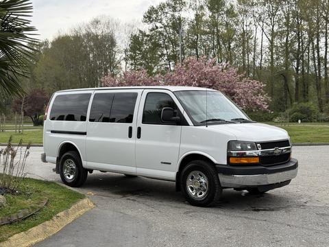 A white 2004 Chevrolet Express van with large windows and chrome wheels parked with greenery in the background