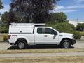 A white 2019 Ford F-150 pickup truck with a utility bed and a ladder rack on top parked on a gravel surface