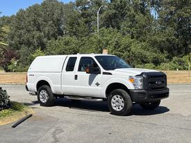 A white 2014 Ford F-250 SD with a truck bed cover parked on a paved area featuring large wheels and four-wheel drive capability