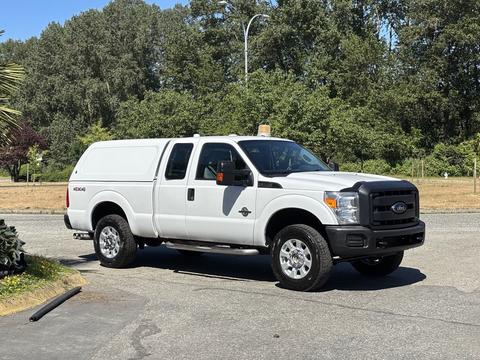 A white 2014 Ford F-250 SD with a truck bed cover parked on a paved area featuring large wheels and four-wheel drive capability