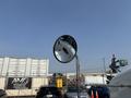A circular side mirror of a 2020 Peterbilt 330 reflecting part of the cabin and a glimpse of vehicles and industrial equipment in the background