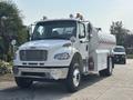 A 2010 Freightliner M2 106 truck in white with an attached tank for liquid transport featuring side mirrors and multiple lights on the roof
