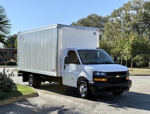 A 2024 Chevrolet Express box truck with a white body and black grille parked in a lot with greenery in the background