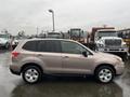 A 2016 Subaru Forester in a light brown color with five doors and a roof rack parked in a lot with large vehicles in the background