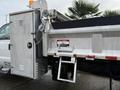 A 2012 Ford F-550 dump truck with a silver body and a metal compartment attached to the driver side featuring warning labels and a hydraulic lift mechanism