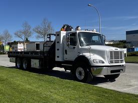 A 2012 Freightliner M2 112 truck with a flatbed and storage compartments is parked with its cab facing forward and three axles visible