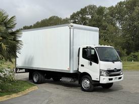 A 2018 Hino 195 truck with a white box cargo area parked on a road with trees in the background