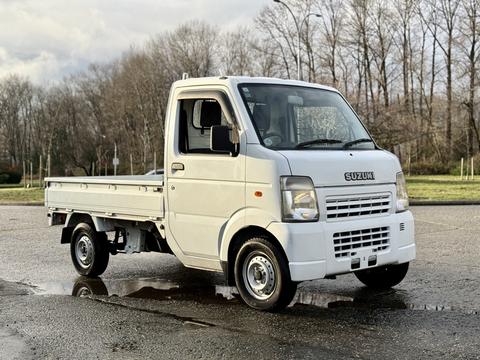 A white 2008 Suzuki Carry pickup truck with a flatbed and a compact design parked on a wet surface