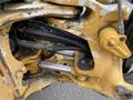Close-up of a yellow hydraulic system on a 2012 John Deere 310SJ showing exposed pipes and mechanical components with signs of wear and rust