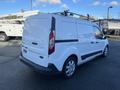 A white 2017 Ford Transit Connect van with sliding side doors and a roof rack viewed from the rear angle