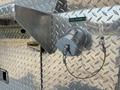 A close-up of a metallic rear discharge fitting on a diamond plate surface with a connected cable and a small green label reading REAR DISCHARGE