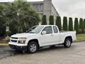 A white 2011 Chevrolet Colorado pickup truck with a two-door cab and a bed is parked on a paved surface with greenery in the background