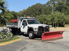 A white 2008 Ford F-450 SD with a snow plow attached is parked in a lot the truck has a flatbed and is equipped for snow removal