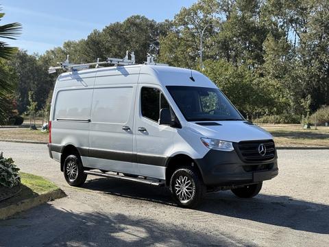 A 2023 Mercedes-Benz Sprinter 2500 van with a white and silver exterior and roof racks parked on a gravel surface