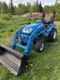 A blue 2021 LS XJ2025H tractor with a front loader attachment sitting on grass with some yellow flowers visible near the front tires