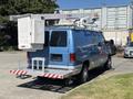 A blue 2008 Ford E-350 utility van with a raised bucket lift attached to the roof and a rear step platform for access