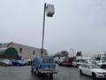 A blue 2006 Ford Econoline van with a raised aerial lift attached to its roof, positioned in a parking area with various vehicles around it