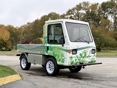 A 2019 CanEV Might-E electric utility vehicle with a white body and green leaf designs on the sides features a cab with large windows and a flatbed for cargo.