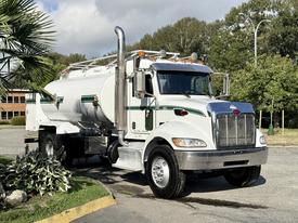 A white 2015 Peterbilt 337 truck with a silver fuel tank and green stripes parked on a pavement with a shiny front grille and large side mirrors