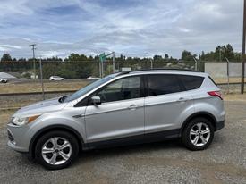 A silver 2015 Ford Escape parked on gravel with its driver side facing the viewer showcasing its sleek design and alloy wheels