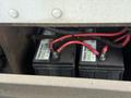 Two black batteries with red cables attached, labeled with "CANADA PROOF MATECH," positioned inside a compartment of a vehicle.