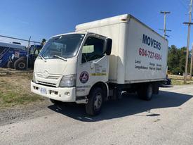 A 2014 Hino 195 box truck with a white cargo area and a company logo on the side parked on a road